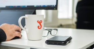 Kuvituskuva: Työpöydällä Solidabis-kahvikuppi, silmölasit ja puhelin. Reunassa näkyy tietokoneen näyttö ja hiirtä käyttävä käsi.